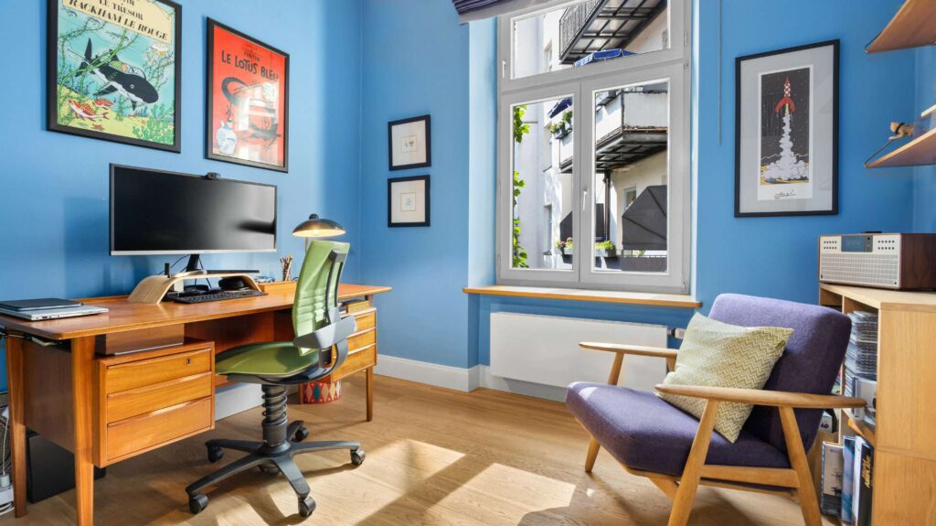 Arbeitszimmer der Altbauwohnung in Haidhausen mit blauen Wänden, Holzschreibtisch, grünem Bürostuhl und Kunstdrucken