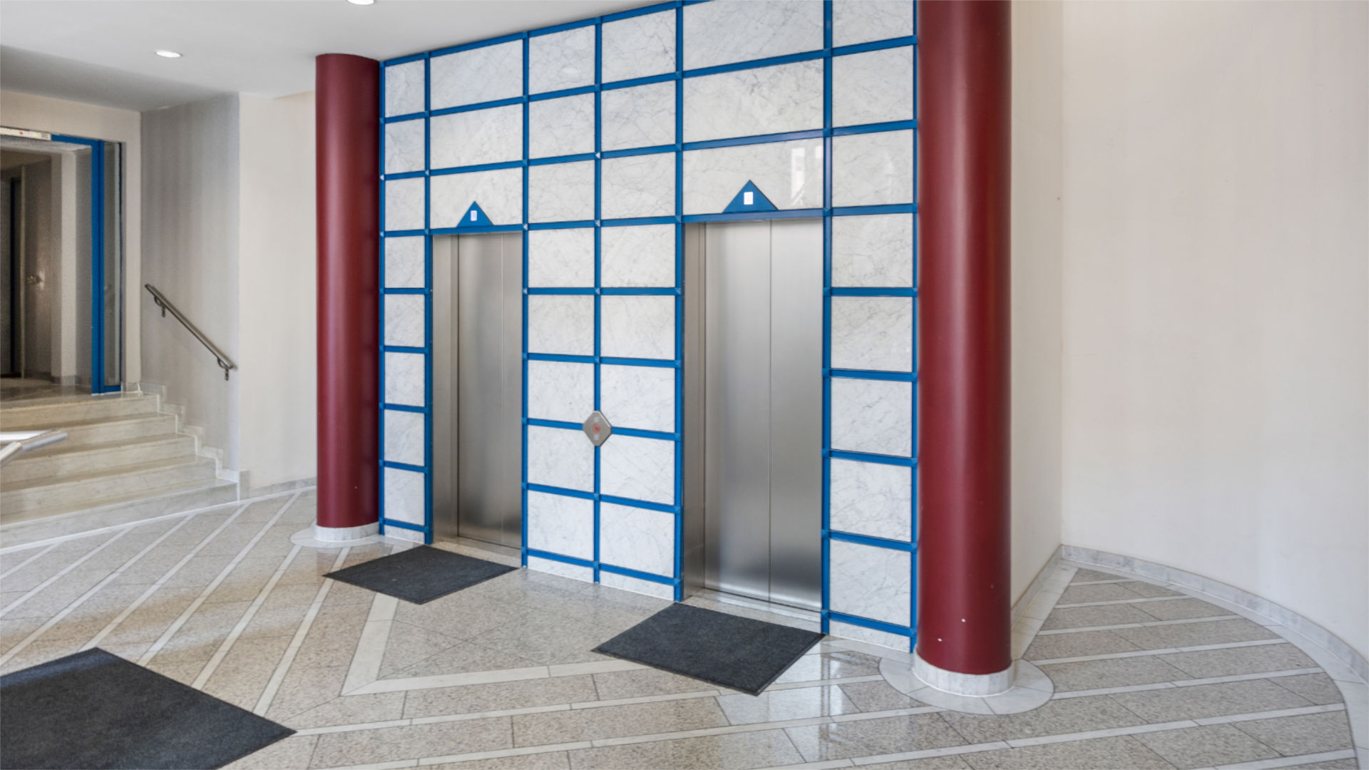 Two elevator doors between red columns and a mosaic wall in the entrance area of an apartment building in Munich-Haidhausen