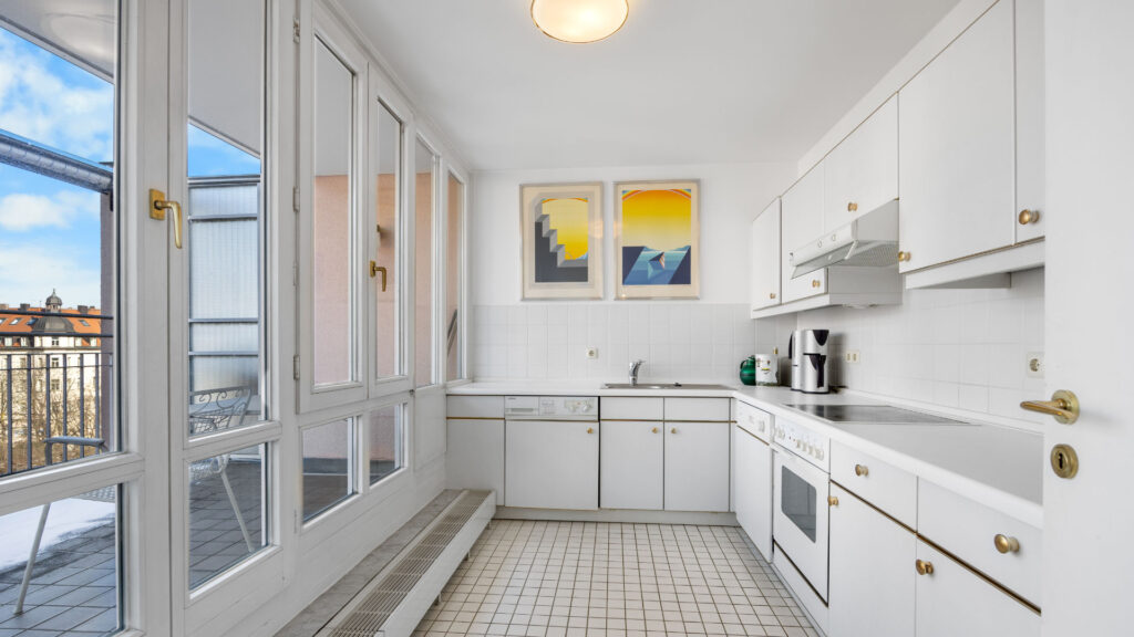 Narrow white kitchen with tiled floor, yellow wall art and access to the glazed balcony in Haidhausen