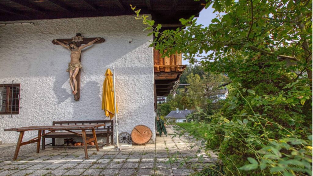 Gepflasterte Terrasse mit Kruzifix an der Fassade und Sitzbank des Hauses in Kochel am See