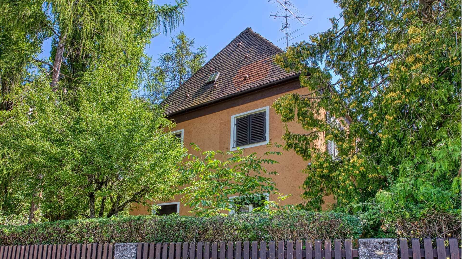 Plastered residential building with a dark tile roof, surrounded by dense greenery on the property in Munich-Laim