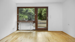 Empty living room with window, patio door, and light hardwood floor in a rented apartment in Munich-Lehel