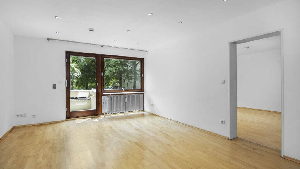 Leeres Wohnzimmer mit dunkler Balkontür, Holzboden und Durchgang zu weiterem Raum in Mietwohnung München-Lehel