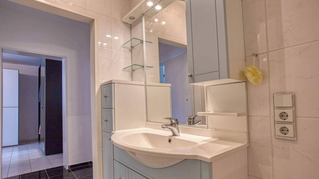 Badezimmer in Maxvorstadt mit blau-grauem Waschtisch, Spiegel mit Glasablagen und cremefarbenen Wandfliesen.
