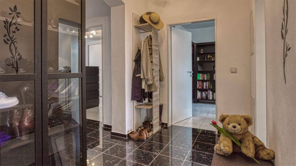 Flur mit dunklem Fliesenboden, Garderobe mit Jacken, Glastür zum Bücherregal und einem Teddybär auf dem Boden.
