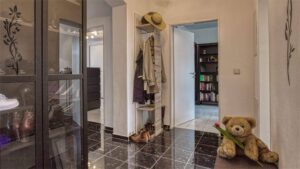 Flur mit dunklem Fliesenboden, Garderobe mit Jacken, Glastür zum Bücherregal und einem Teddybär auf dem Boden.