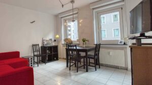 Wohnzimmer in Maxvorstadt mit rotem Sofa, dunklem Holztisch, mehreren Stühlen und Fensterblick auf Nachbargebäude.
