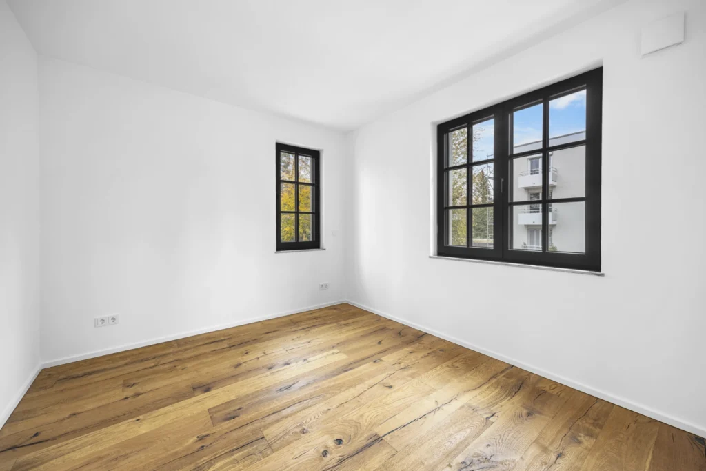 Empty bedroom with wooden floorboards and two windows in a rental apartment in Pöcking