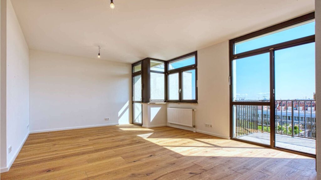 Leerer Wohnraum in Sendling mit großem Panoramafenster, Parkettboden und Blick auf die Nachbarschaft.