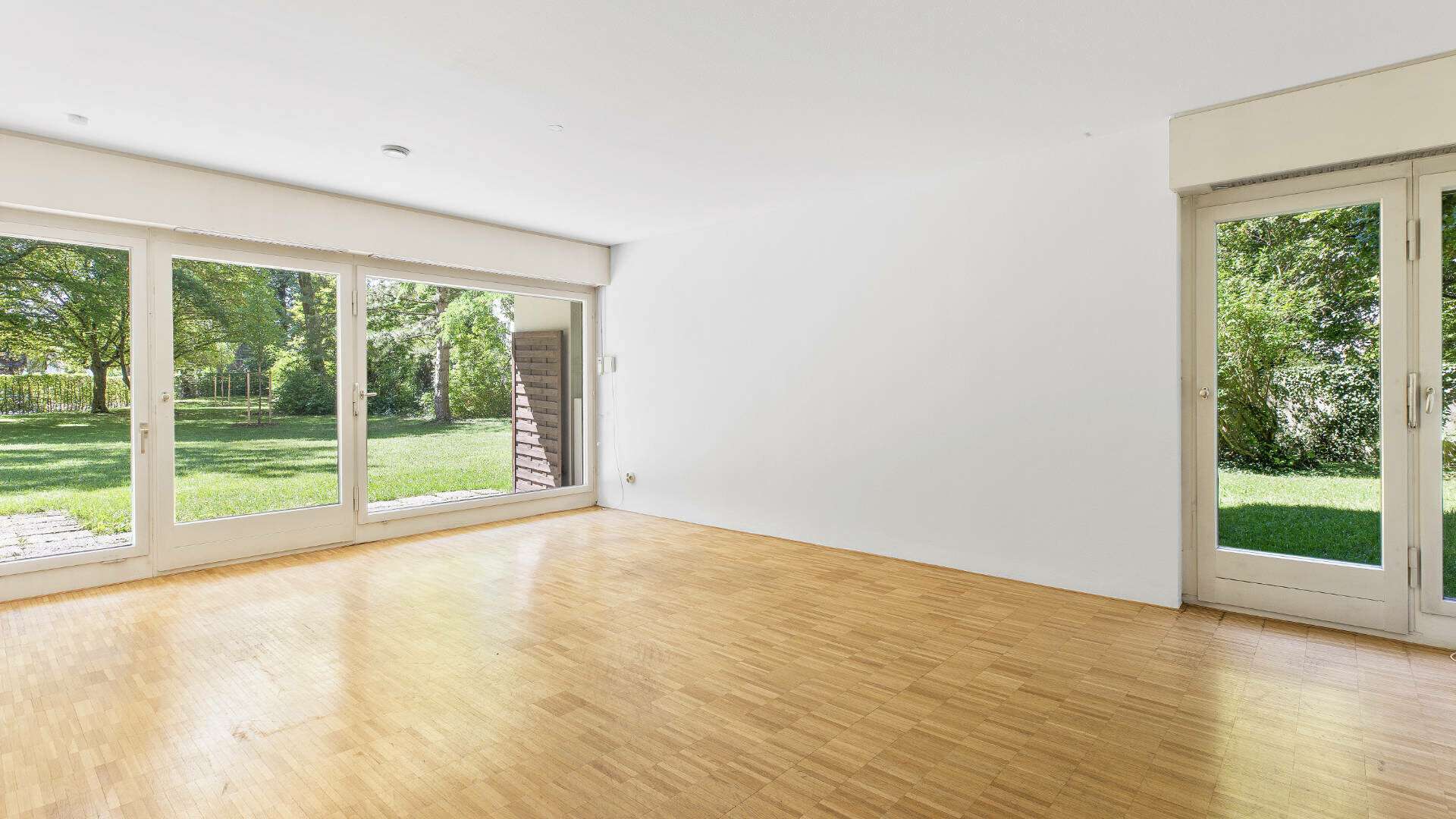 Leeres Wohnzimmer in Solln mit zwei Terrassenzugängen, Parkettboden und Gartenblick.