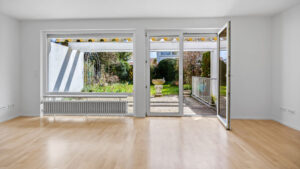 Empty living room with large front window and open patio door to the garden in an end-of-terrace house in Unterhaching