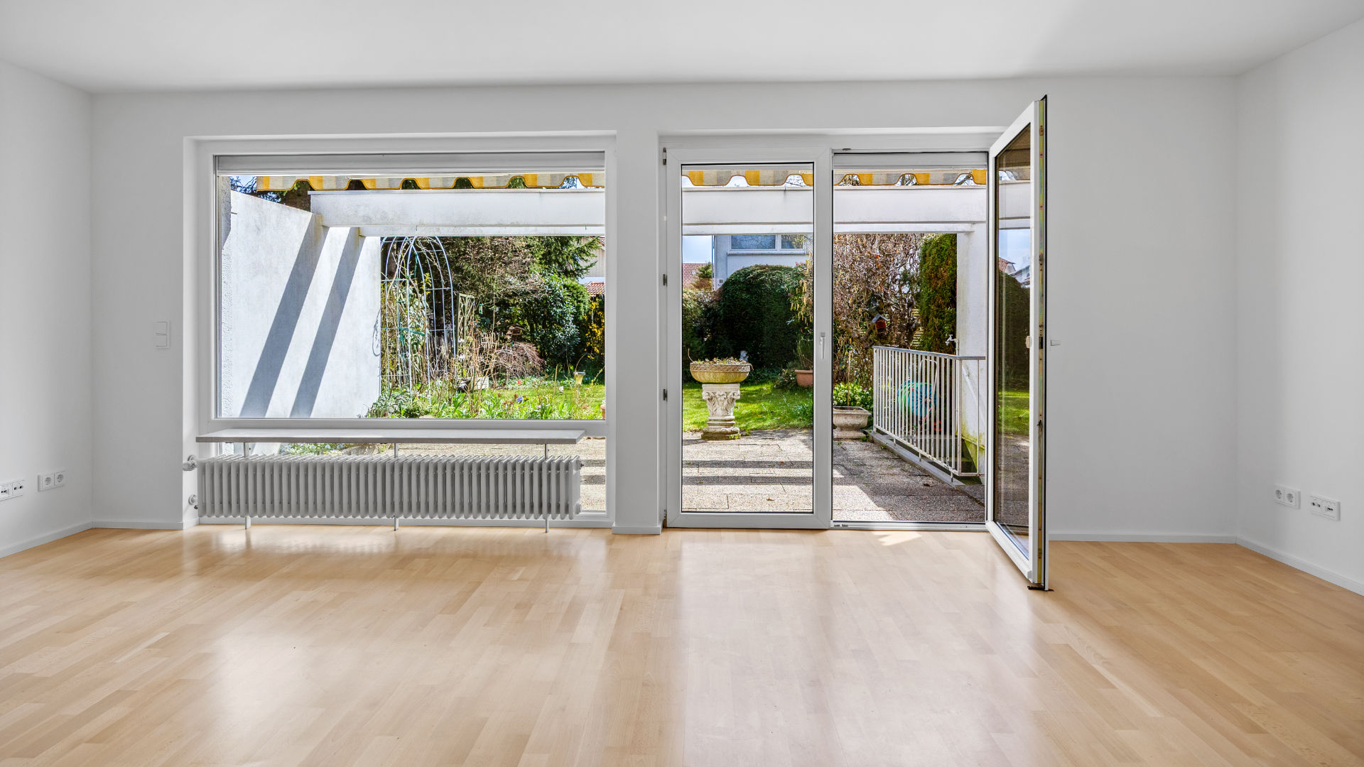 Empty living room with large front window and open patio door to the garden in an end-of-terrace house in Unterhaching