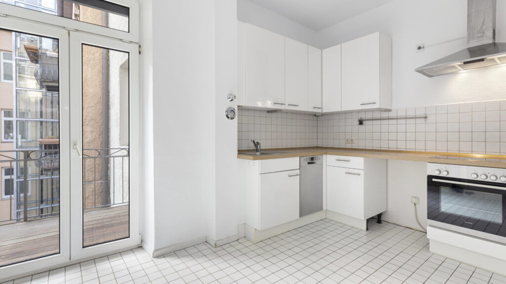 Older eat-in kitchen with white cabinets, balcony door, and tile floor in an old-style apartment in the Gärtnerplatz district of Munich