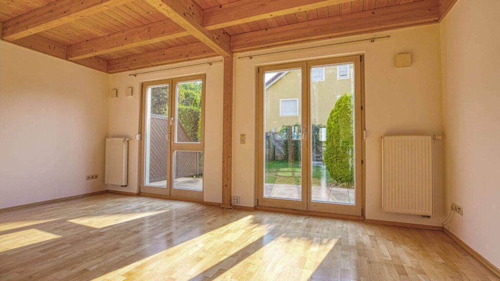 Leeres Wohnzimmer des Hauses in Germering mit sichtbaren Holzbalken, zwei Terrassentüren und Blick in den Garten