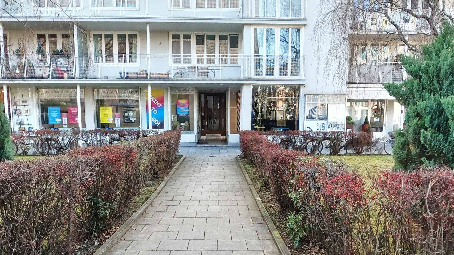 Paved path between hedges leading to the facade of a building with shop windows in Munich-Maxvorstadt