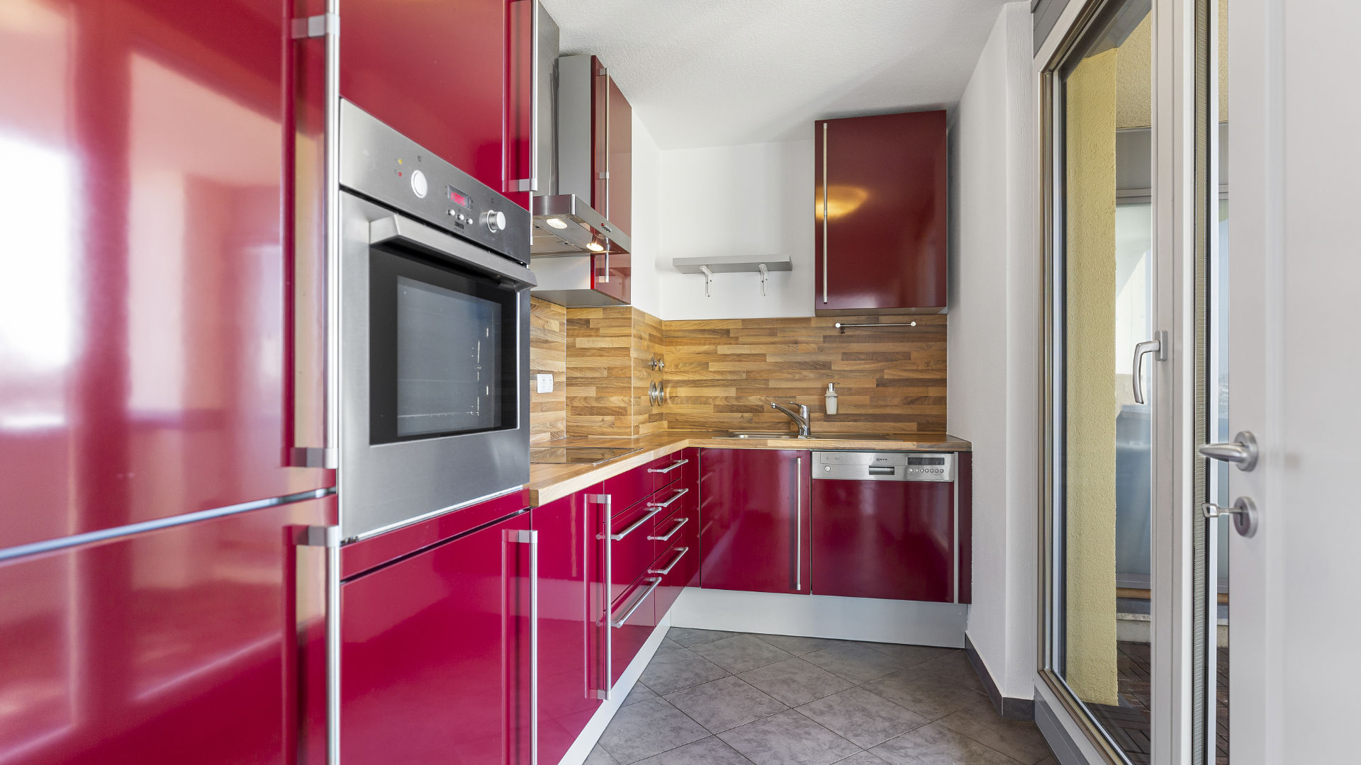 Einbauküche mit glänzend roten Fronten, Einbaubackofen und Balkonzugang in einer Wohnung in München-Milbertshofen