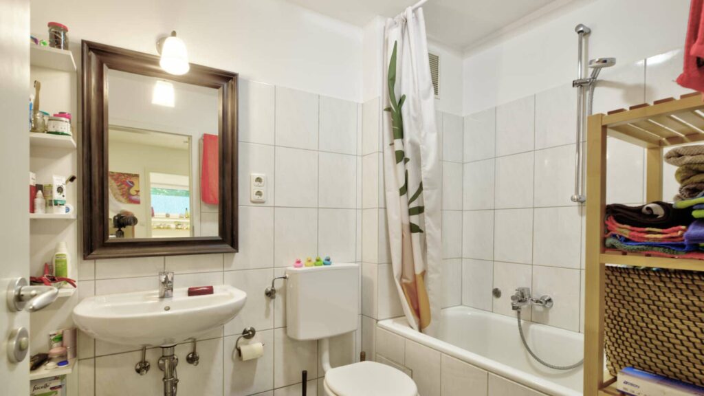Small, older bathroom with mirror, rattan shelf, bathtub and patterned shower curtain in Perlach