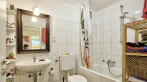 Small, older bathroom with mirror, rattan shelf, bathtub and patterned shower curtain in Perlach
