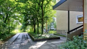 Paved path through trees leads to a house entrance with stairs and a parked bicycle in Perlach