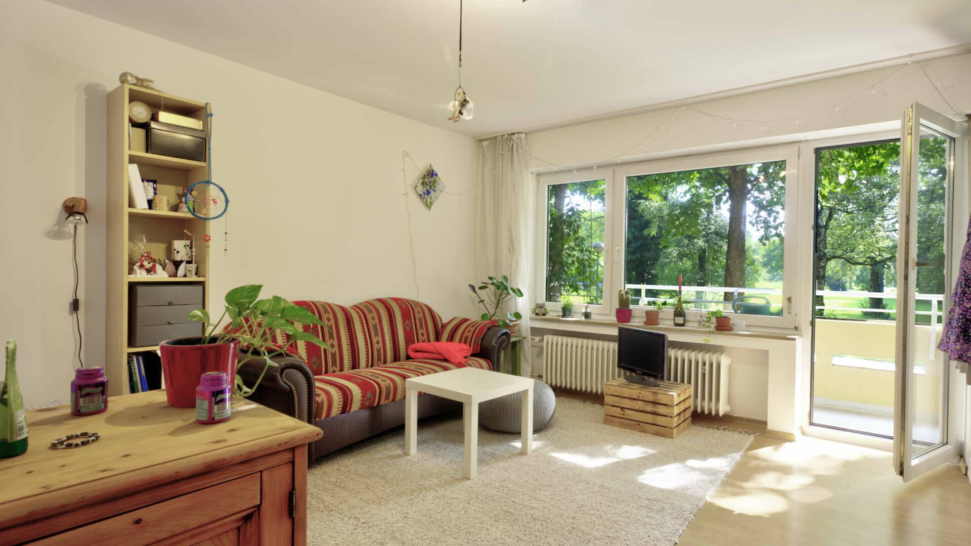 Wohnzimmer mit gemütlicher Sitzecke, Bücherregal und Balkontür mit Blick auf eine Grünfläche in Perlach