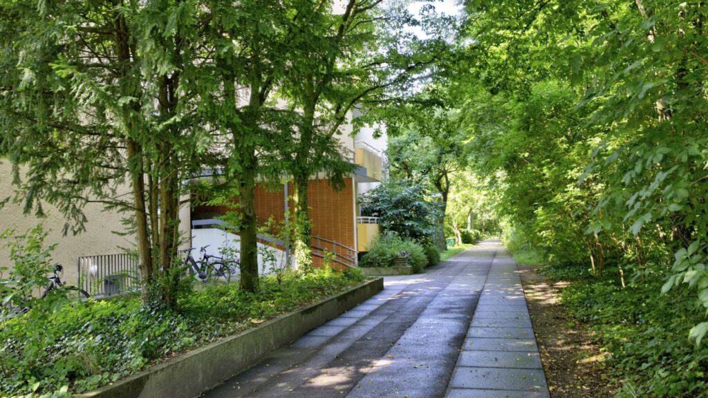 Paved path between trees leads to a building with orange-colored window shutters in Munich-Perlach