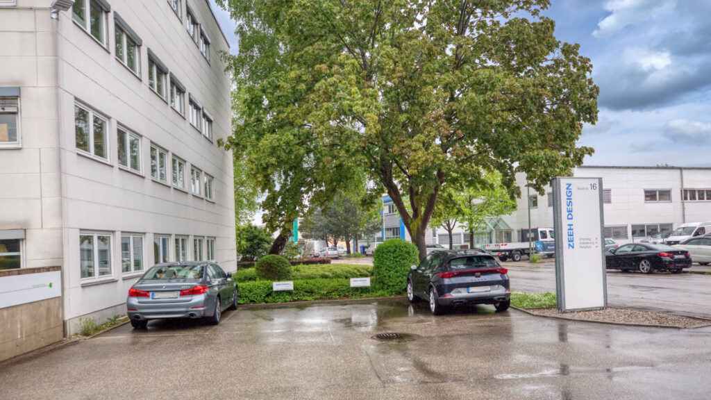 Building corner with tree, advertising pylon, and parking spaces at the Puchheim office building – Rogers Immobilien