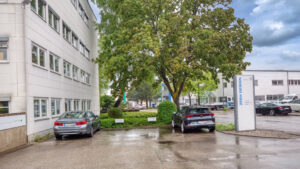 Building corner with tree, advertising pylon, and parking spaces at the Puchheim office building – Rogers Immobilien