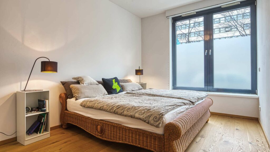 Bedroom with rattan bed, nightstand and floor lamp by the window in the apartment in Schwabing – Rogers Immobilien