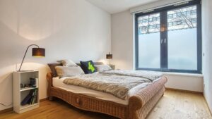 Bedroom with rattan bed, nightstand and floor lamp by the window in the apartment in Schwabing – Rogers Immobilien
