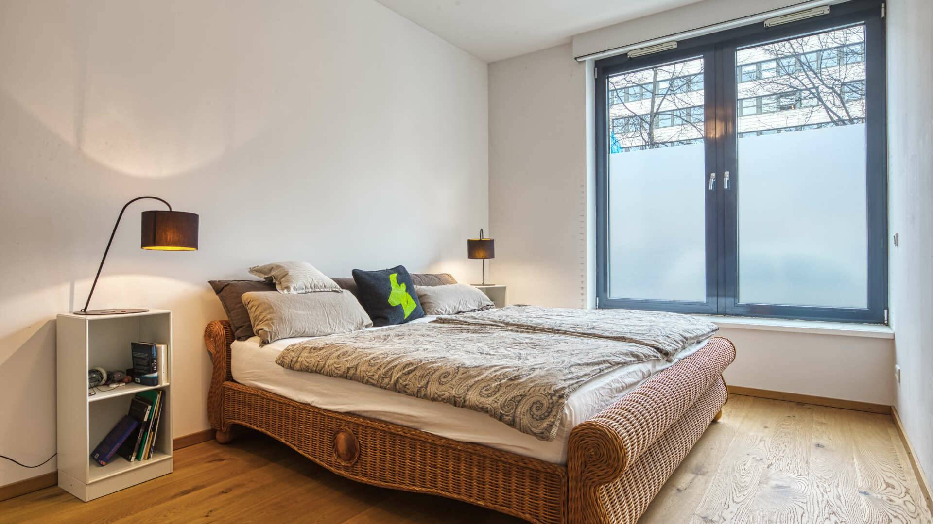 Bedroom with rattan bed, nightstand and floor lamp by the window in the apartment in Schwabing – Rogers Immobilien