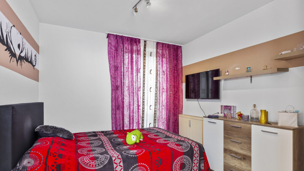 Bedroom with red duvet cover, TV and small kitchenette in an apartment in Munich-Untermenzing