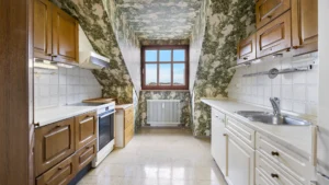 Unrenovated kitchen under the slanted ceiling with wooden cabinets and old wallpaper in Munich-Berg am Laim – Rogers Immobilien