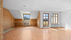 Empty living space with two windows, patio door, and wooden floor before renovation in Munich Berg am Laim – Rogers Immobilien