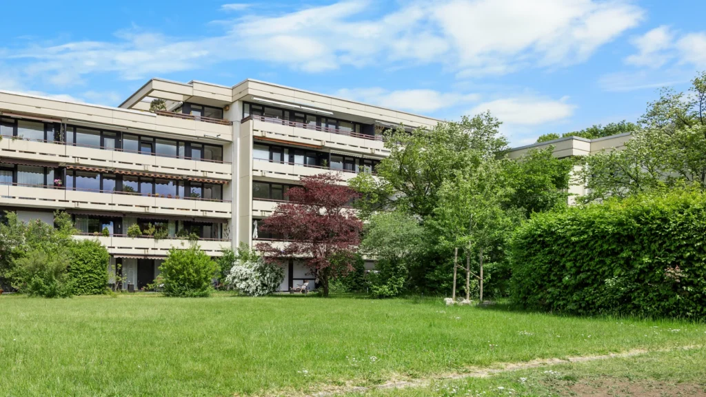 Blick durch Bäume auf das mehrstöckige Wohngebäude der Anlage in München-Forstenried mit Grünfläche
