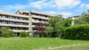 Blick durch Bäume auf das mehrstöckige Wohngebäude der Anlage in München-Forstenried mit Grünfläche