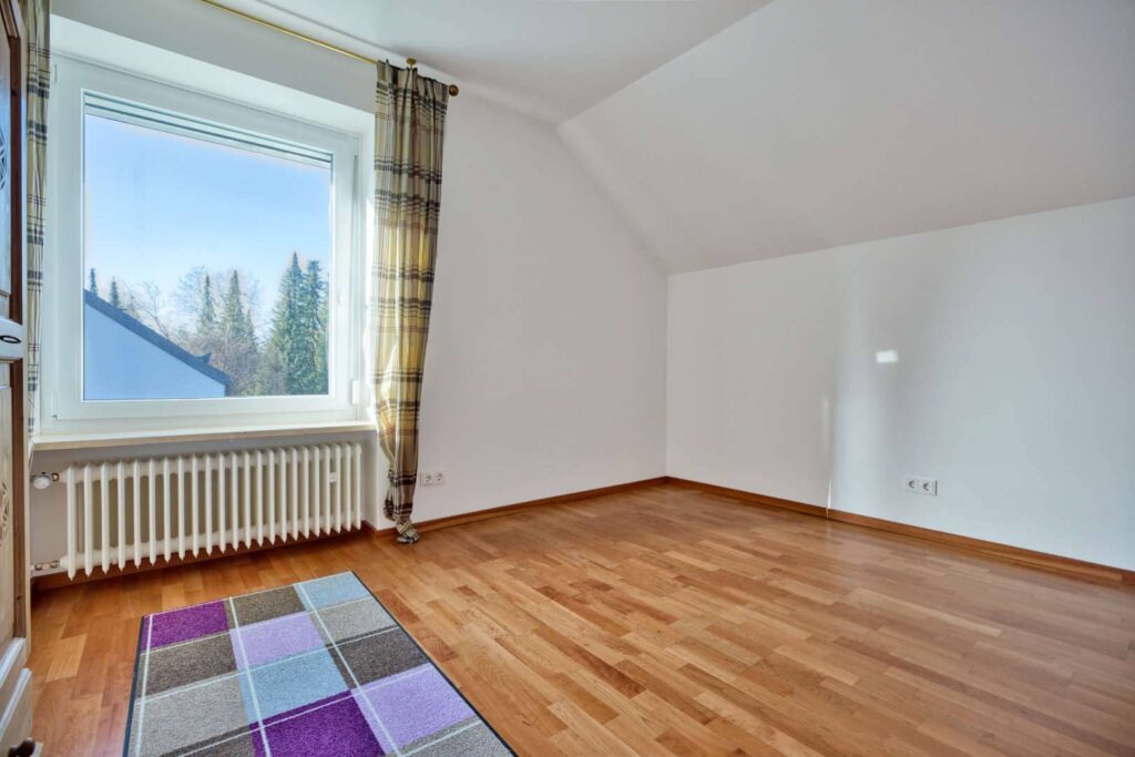 Empty attic room with a colorful carpet and window view of trees in Munich-Obermenzing – Rogers Immobilien